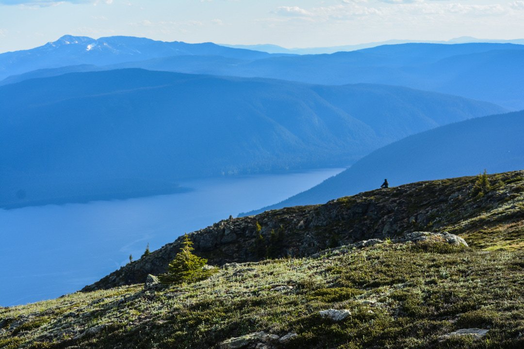 man sitting peacefully on top of mountain with deep blue hues, mountains and lake in background