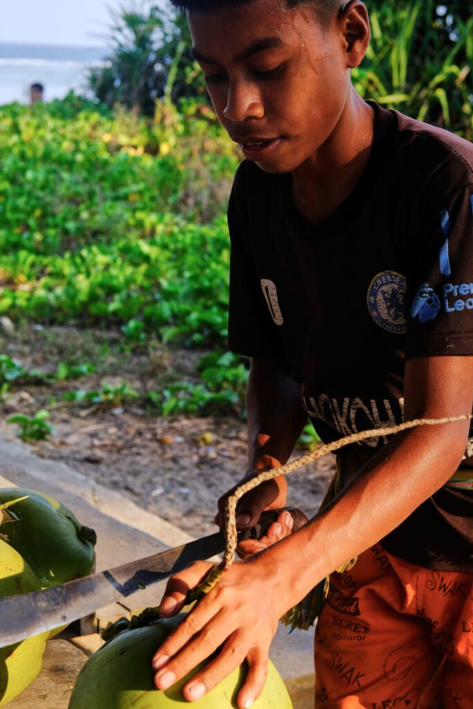 A local village boy by the name of Sepri carefully slices open a coconut with his machete as sweat runs down his face