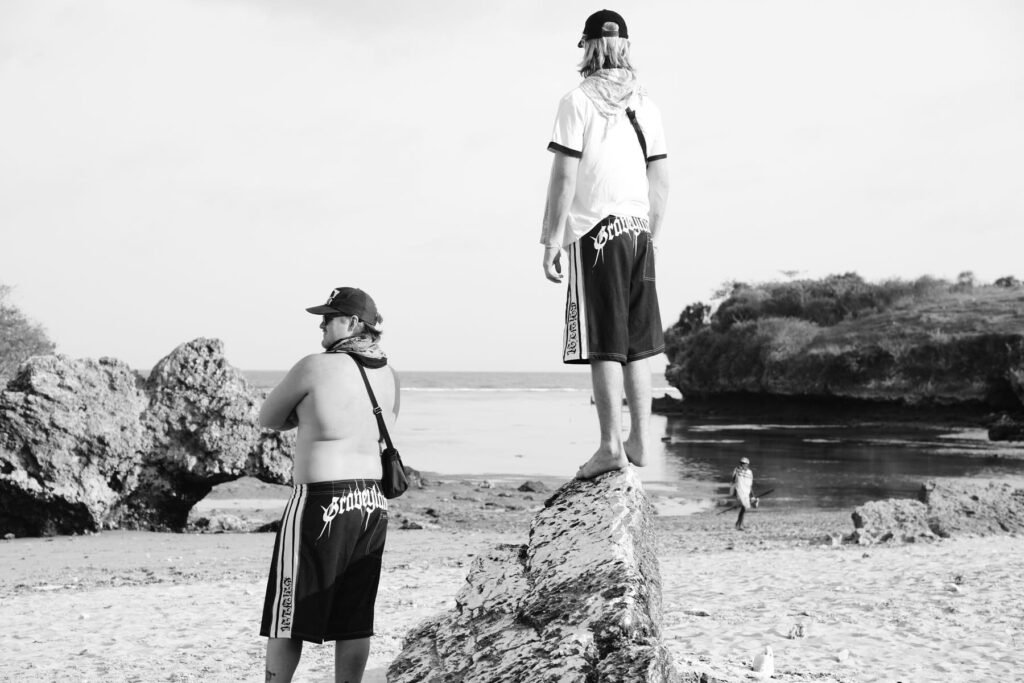 Boardies, hats and t-shirts on. One guy standing on top of rock staring out to sea, the other looking out at the horses near the beach