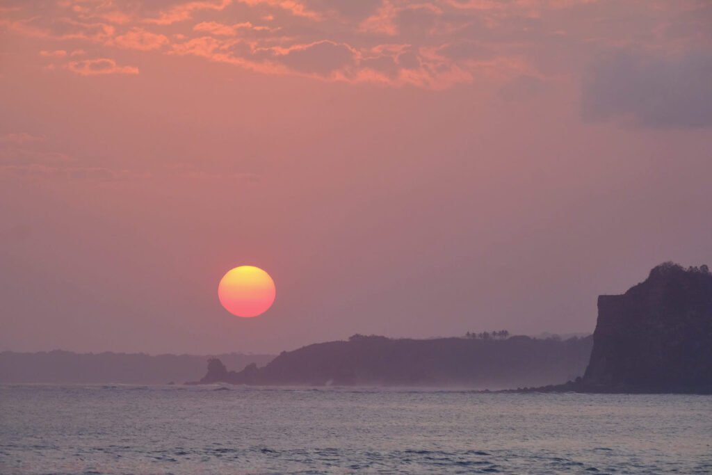 The sun set is big and red as it slowly drops down to the horizon next to some cliffs.