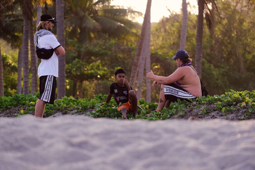 Two of the Australian boys stand and sit either side of the local villager Sepri who cut open some coconuts for us. The sun sets behind the beach here.