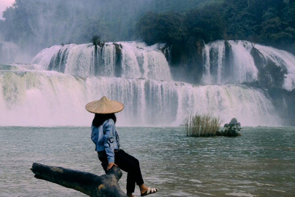 Women sits on a tree trunk looking out to the main cascade of Ban Gioc waterfall. Wearing a traditional Vietnamese hat.