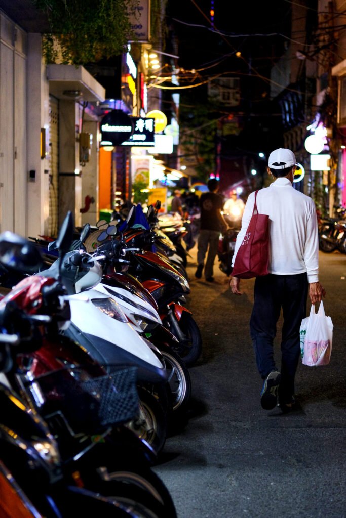 Scooters line the dimly lit streets of Hanoi. A man is walking with his food in a plastic bag,