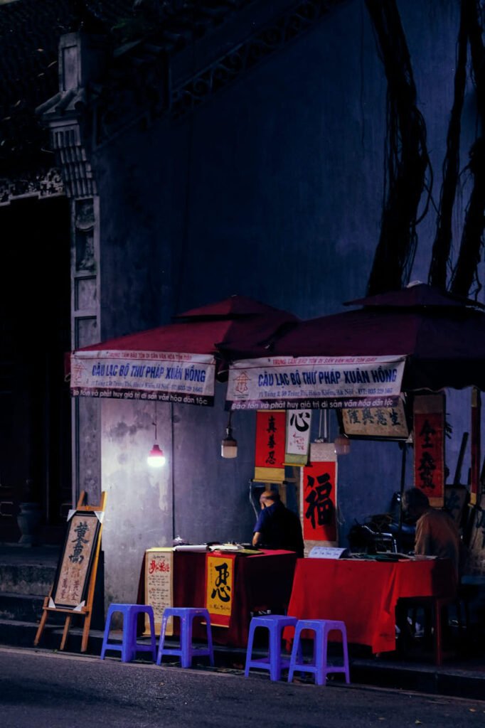 A man sits in his temporary roadside shelter, with calligraphy paintings as signage. Hanoi, Vietnam.