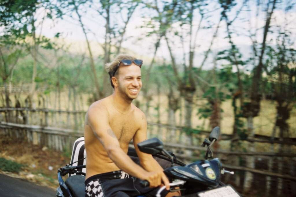 A man drives shirtless through the back roads on a surf trip, smiling and free