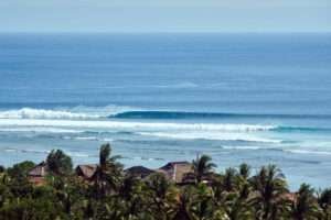 A perfect left hand wave peels along the reef of Desert Point. Vantage point from above. The water is deep blue and the vibe tropical with coconut trees in the foreground.