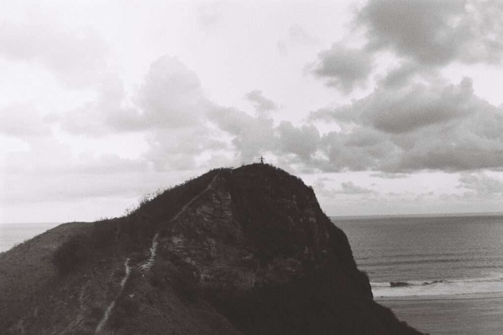 A man stands on top of the hill with arms raised in the distance overlooking the ocean, Kuta Lombok