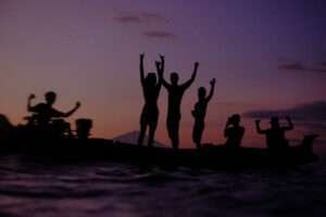 The sunsets on a magical day on the water. Oranges and purples splay across the sky as surfers and boat crew are stoked standing with their arms up in joy